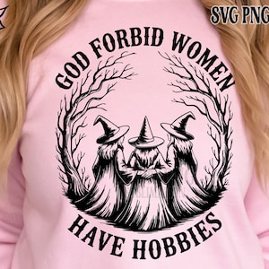 May include: A light pink sweatshirt featuring a black graphic of three witches in pointed hats, surrounded by a tree-like design. The text reads "GOD FORBID WOMEN HAVE HOBBIES" in a circular arrangement. The design is in the center of the sweatshirt.