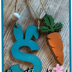 May include: Two wooden Easter ornaments, one shaped like a bunny with blue paint and the other shaped like a carrot with green leaves and orange paint. The ornaments are hanging from twine and have a white bead at the top. The text "On The Beach Boutique" is visible at the bottom of the image.