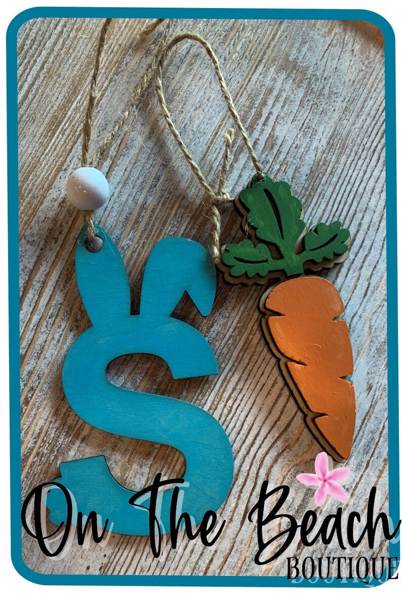 May include: Two wooden Easter ornaments, one shaped like a bunny with blue paint and the other shaped like a carrot with green leaves and orange paint. The ornaments are hanging from twine and have a white bead at the top. The text "On The Beach Boutique" is visible at the bottom of the image.