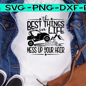May include: White t-shirt with the text "the BEST THINGS in LIFE MESS UP YOUR HAIR" in black. The design includes a side-by-side vehicle, mountains, and a log. The shirt is displayed on a denim jacket and a brown purse.