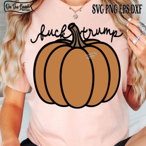May include: A light pink t-shirt featuring a large brown pumpkin graphic with the words "fuck trump" in black script. The shirt also has the text "SVG PNG EPS DXF" in the upper right corner.