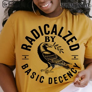 May include: Mustard yellow t-shirt with a black graphic of a crow holding a sprig, encircled by the words "RADICALIZED BY BASIC DECENCY." The design is centered on the shirt. The shirt is made of a soft material.