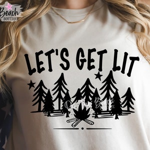 以下が含まれることがあります： 黒い文字で「Let's Get Lit」と書かれた白いTシャツ。焚き火と松の木のグラフィックが描かれている。