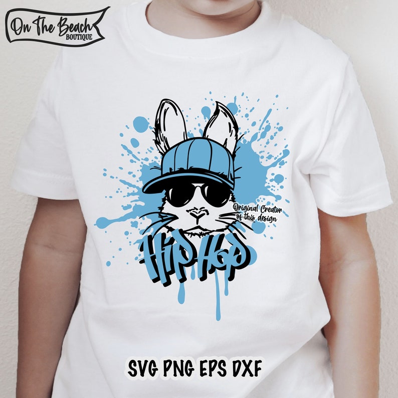 K&ouml;nnte beinhalten: Ein wei&szlig;es T-Shirt mit einer blauen und schwarzen Grafik eines Kaninchens, das eine Sonnenbrille und einen Hut tr&auml;gt. Das Kaninchen ist von blauen Farbflecken umgeben und das Wort "Hip Hop" ist darunter geschrieben. Der Text "Original Creator of this design" ist ebenfalls enthalten.