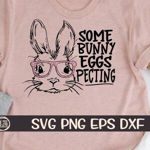 Puede incluir: Una camiseta rosa con una ilustración en blanco y negro de un conejito con gafas. El texto "Some Bunny Eggs-pecting" está escrito en negro en la camiseta.