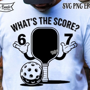 Pode incluir: Camiseta branca com um design gráfico preto com uma raquete de pickleball, uma bola e o texto "WHAT'S THE SCORE?" com os números 6 e 7. A raquete tem mãos de desenho animado.
