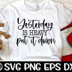 Könnte beinhalten: Weißes T-Shirt mit dem Text "Yesterday is heavy put it down" in schwarzer Kursivschrift. Das Shirt liegt auf einer Holzoberfläche mit roten High Heels und Weinkorken.