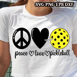 May include: White t-shirt with a black peace sign, a black heart, and a yellow pickleball. The text "peace love pickleball" is printed below the symbols.