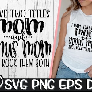 May include: A white tank top with black text that reads "I have two titles mom and... bonus mom and I rock them both". The text is in a bold, cursive font. The tank top is being worn by a person with long brown hair. The background is a white wooden surface.