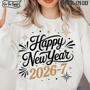 May include: White sweatshirt with the text "Happy New Year 2026-7" in black and gold. The design includes starburst and dot accents. The sweatshirt has the "On The Beach Boutique" logo in the upper left corner.
