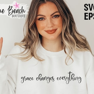 May include: A white sweatshirt with the text "grace changes everything" in black cursive lettering. The model is wearing the sweatshirt and giving a peace sign.