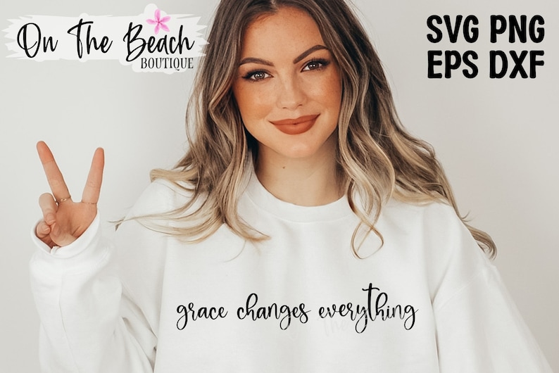 May include: A white sweatshirt with the text "grace changes everything" in black cursive lettering. The model is wearing the sweatshirt and giving a peace sign.