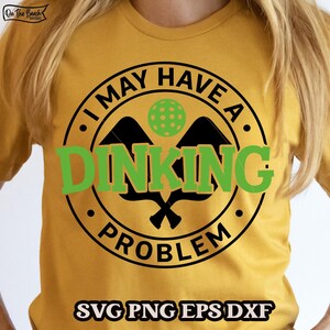 Peut inclure: T-shirt jaune moutarde avec un motif noir et vert. Le motif indique "I MAY HAVE A DINKING PROBLEM" dans un cercle avec des raquettes et une balle de pickleball. Le bas du t-shirt porte l'inscription "SVG PNG EPS DXF".