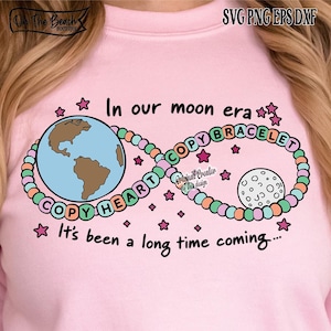 Op de afbeelding: Roze sweatshirt met een grafisch ontwerp met de aarde, maan en een oneindigheidssymbool gemaakt van kralen. Tekst bevat "In our moon era" en "It's been a long time coming..."