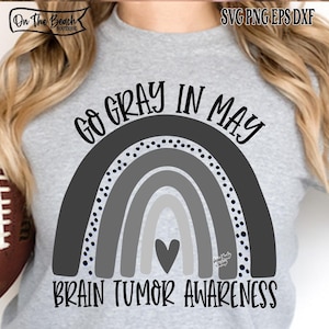 Könnte beinhalten: Ein graues T-Shirt mit einem Regenbogen-Design in Grautönen, einem Herz und dem Text "GO GRAY IN MAY BRAIN TUMOR AWARENESS". Das Shirt hat auch den Text "On The Beach Boutique" oben.