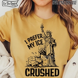 Pode incluir: Camiseta amarelo mostarda com um gráfico preto da Estátua da Liberdade segurando uma tocha, em pé sobre um bloco de gelo. O texto "I PREFER MY ICE CRUSHED" está impresso abaixo.