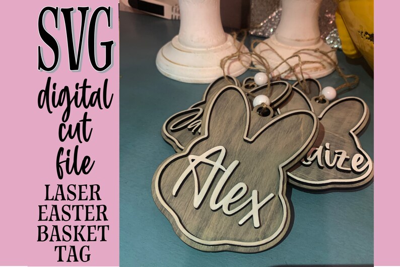 May include: A set of laser cut wooden bunny-shaped Easter basket tags with names "Alex" and "Arize" written on them. The tags are brown with a white woodgrain pattern and have a white bead attached to a twine string.