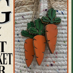 May include: Three wooden carrot ornaments with orange bodies and green leafy tops hang against a woven background. The carrots are suspended by twine. The image is framed with a green border.