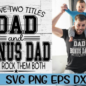 May include: A black and white graphic design with the text "I have two titles Dad and Bonus Dad and I rock them both". The graphic is on a white wood background. A man is holding a child on his shoulders. The child is wearing a blue shirt. The man is wearing a black t-shirt with the same graphic design in white.