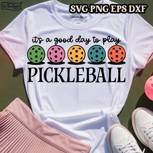 May include: White t-shirt with the text "it's a good day to play PICKLEBALL" in black. Five colorful pickleball paddles are arranged in a row above the text.