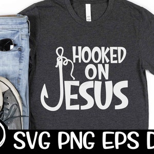 May include: A dark gray t-shirt with a white hook and the text "Hooked on Jesus" printed on the front. The shirt is displayed on a white background with a pair of jeans, sunglasses, and sneakers.