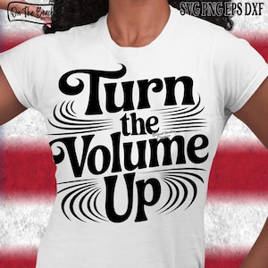 Peut inclure: T-shirt blanc avec l'inscription "Turn the Volume Up" en lettres noires et grasses. Le motif comprend des lignes stylisées en forme de vagues au-dessus et en dessous du texte. Le t-shirt est de style col rond.