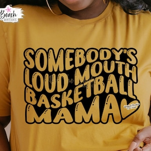 May include: A yellow t-shirt with black text that reads "Somebody's Loud Mouth Basketball Mama" with a small heart graphic that says "Original Designs This Shop".