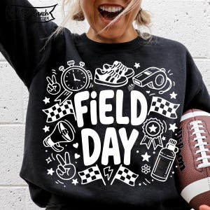 K&ouml;nnte beinhalten: Schwarzes Sweatshirt mit wei&szlig;en Grafikelementen, darunter eine Stoppuhr, Turnschuhe, eine Trillerpfeife, karierte Flaggen und die Worte "FIELD DAY". Ein brauner Football wird im Bild gehalten.