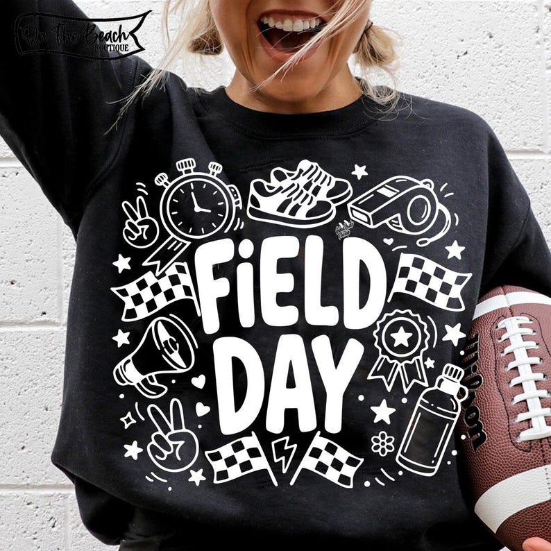 K&ouml;nnte beinhalten: Schwarzes Sweatshirt mit wei&szlig;en Grafikelementen, darunter eine Stoppuhr, Turnschuhe, eine Trillerpfeife, karierte Flaggen und die Worte "FIELD DAY". Ein brauner Football wird im Bild gehalten.
