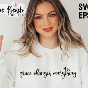 May include: A white sweatshirt with the text "grace changes everything" in black cursive lettering. The model is wearing the sweatshirt and giving a peace sign.
