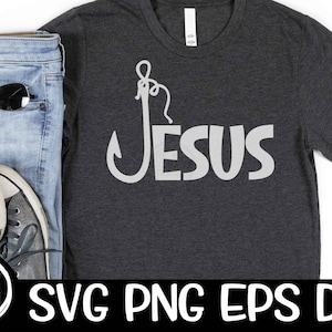 Puede incluir: Una camiseta gris oscuro con un anzuelo de pesca blanco y la palabra "JESUS" impresa en ella. La camiseta está sobre un fondo blanco con un par de jeans, gafas de sol y zapatillas.