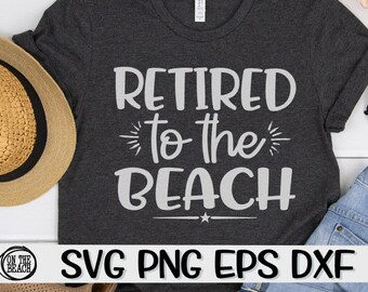 Puede incluir: Una camiseta gris oscuro con el texto "Retired to the Beach" en blanco. La camiseta está sobre una superficie blanca con un sombrero de paja y unos vaqueros azules.