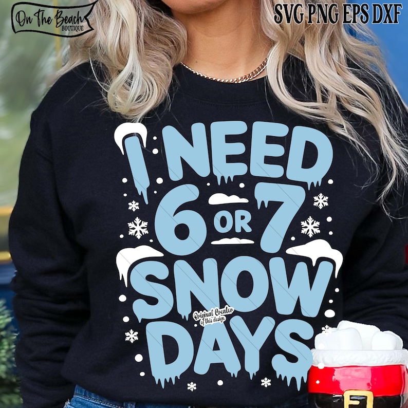 May include: A navy blue sweatshirt featuring the text "I NEED 6 OR 7 SNOW DAYS" in light blue, with snowflake and icicle details. The design includes the "On The Beach Boutique" logo.
