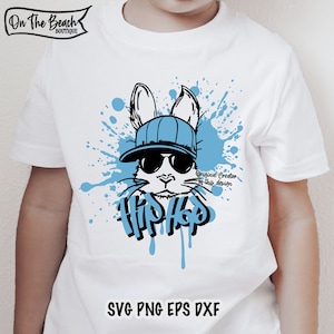 K&ouml;nnte beinhalten: Ein wei&szlig;es T-Shirt mit einer blauen und schwarzen Grafik eines Kaninchens, das eine Sonnenbrille und einen Hut tr&auml;gt. Das Kaninchen ist von blauen Farbflecken umgeben und das Wort "Hip Hop" ist darunter geschrieben. Der Text "Original Creator of this design" ist ebenfalls enthalten.