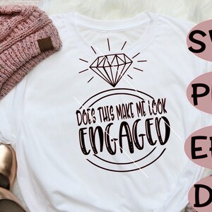 May include: White t-shirt with a black diamond ring graphic and the text "Does this make me look engaged?" The shirt is lying on a white fluffy surface with a pink knitted hat and gold shoes.
