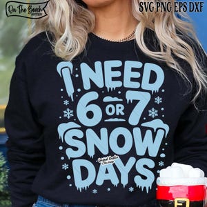 May include: A navy blue sweatshirt featuring the text "I NEED 6 OR 7 SNOW DAYS" in light blue, with snowflake and icicle details. The design uses a playful font. The sweatshirt also has the text "On The Beach Boutique" at the top left.