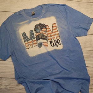 May include: Light blue t-shirt with a bleached white square design. The design features the text "MOM life" with the letters "MOM" in a red, white, and blue American flag pattern. A woman's silhouette is in the center of the design.