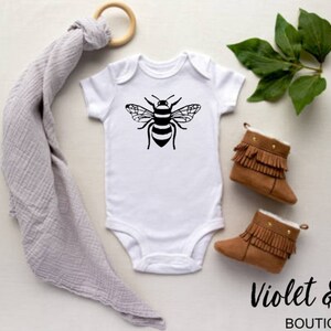 May include: White baby onesie with a black bee graphic. The onesie is on a white surface with a gray swaddle blanket, brown booties, and green leaves.