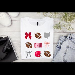 May include: White t-shirt with a design featuring footballs, bows, a red helmet, and the word "OHIO" in red. The shirt is folded on a wooden surface, with a pair of white sneakers, headphones, and a notebook visible.