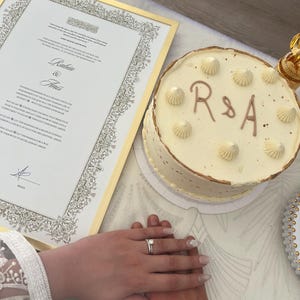 May include: A round white frosted cake with the letters R & A in brown icing and piped cream dollops on top, positioned next to a framed marriage certificate with gold trim featuring the names Rabia and Anis and a signature at the bottom.
