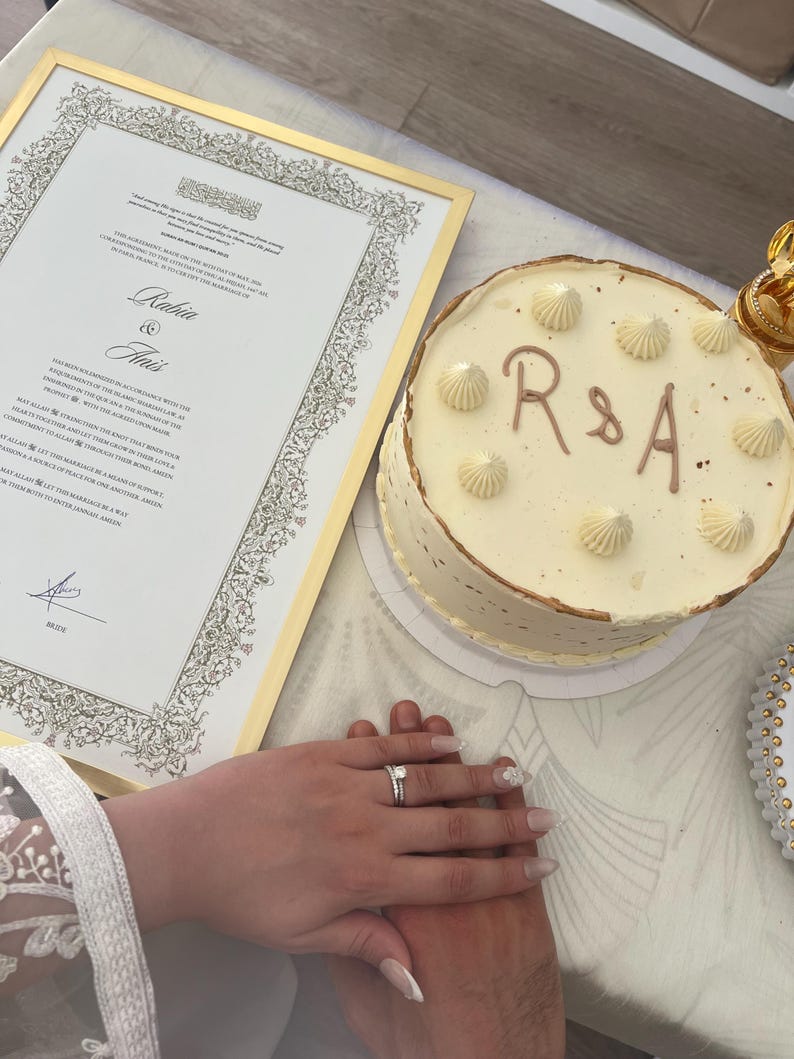 May include: A round white frosted cake with the letters R & A in brown icing and piped cream dollops on top, positioned next to a framed marriage certificate with gold trim featuring the names Rabia and Anis and a signature at the bottom.