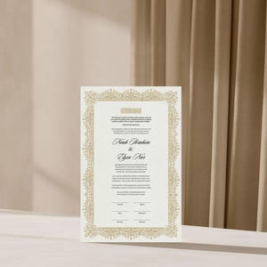 May include: A rectangular wedding invitation card featuring a detailed gold filigree border on a textured white background. The text includes the names Noah Ibrahim and Elyza Nur in a black script font, with additional printed details and lines.