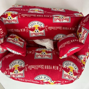 May include: A red fabric tissue box cover featuring the New York Red Bulls logo and team name. The cover has a sofa-like design with two small pillows and armrests. The fabric is patterned with the team's logo.