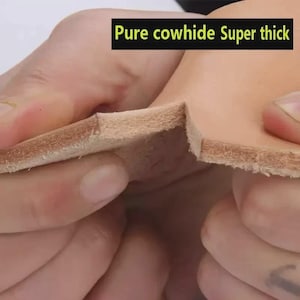 May include: A thick piece of light tan cowhide leather with a visible fibrous texture on the cut edge. The leather is held to display its thickness and density. A black rectangular label with yellow text reads Pure cowhide Super thick.