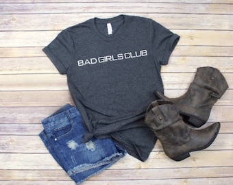 May include: A dark gray t-shirt with the text "BAD GIRLS CLUB" in white lettering. The shirt is paired with denim shorts and brown cowboy boots.