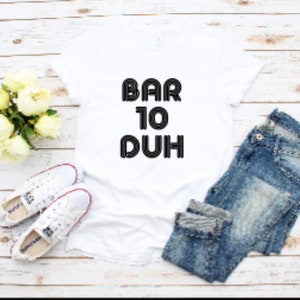 May include: A white t-shirt with the text "BAR 10 DUH" in black, paired with blue jeans and white sneakers. A bouquet of white flowers is also present, creating a casual, everyday look.