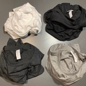 Puede incluir: Cuatro camisetas dobladas en diferentes colores: blanco, negro, gris oscuro y gris claro. Cada camiseta tiene una etiqueta blanca con la palabra "Canvas" impresa en ella.