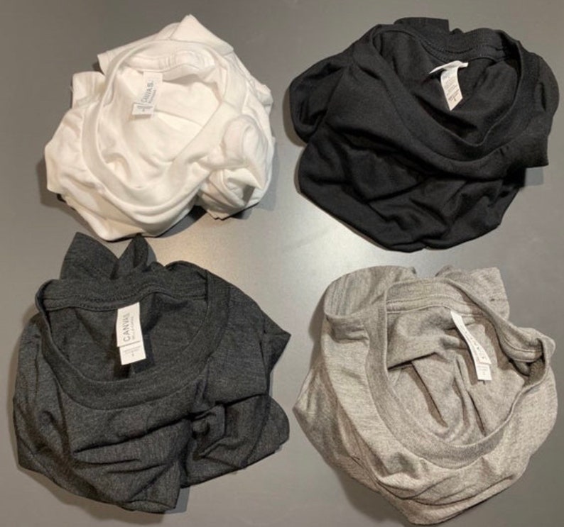 Puede incluir: Cuatro camisetas dobladas en diferentes colores: blanco, negro, gris oscuro y gris claro. Cada camiseta tiene una etiqueta blanca con la palabra "Canvas" impresa en ella.