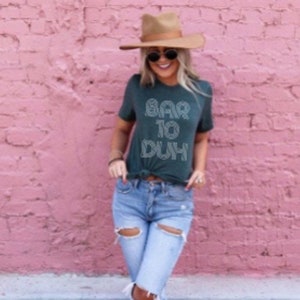 May include: A dark green t-shirt with the text "BAR TO DUH" in white, paired with light blue ripped jeans and a brown hat. The outfit is set against a pink brick wall, creating a casual, stylish look.