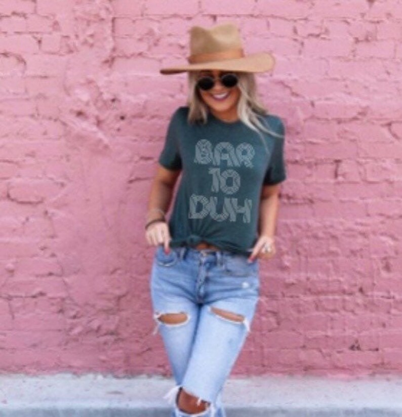 May include: A dark green t-shirt with the text "BAR TO DUH" in white, paired with light blue ripped jeans and a brown hat. The outfit is set against a pink brick wall, creating a casual, stylish look.